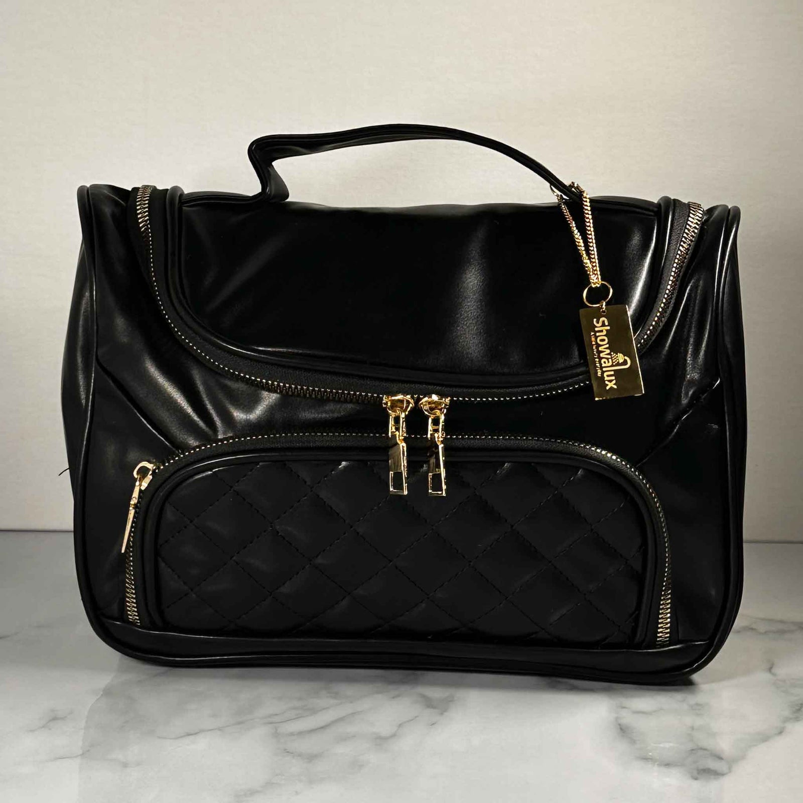 A black handbag style toiletry bag with duel zips for the main section, a side pouch and a gold tag with the showalux logo on a gold chain, on a marble top