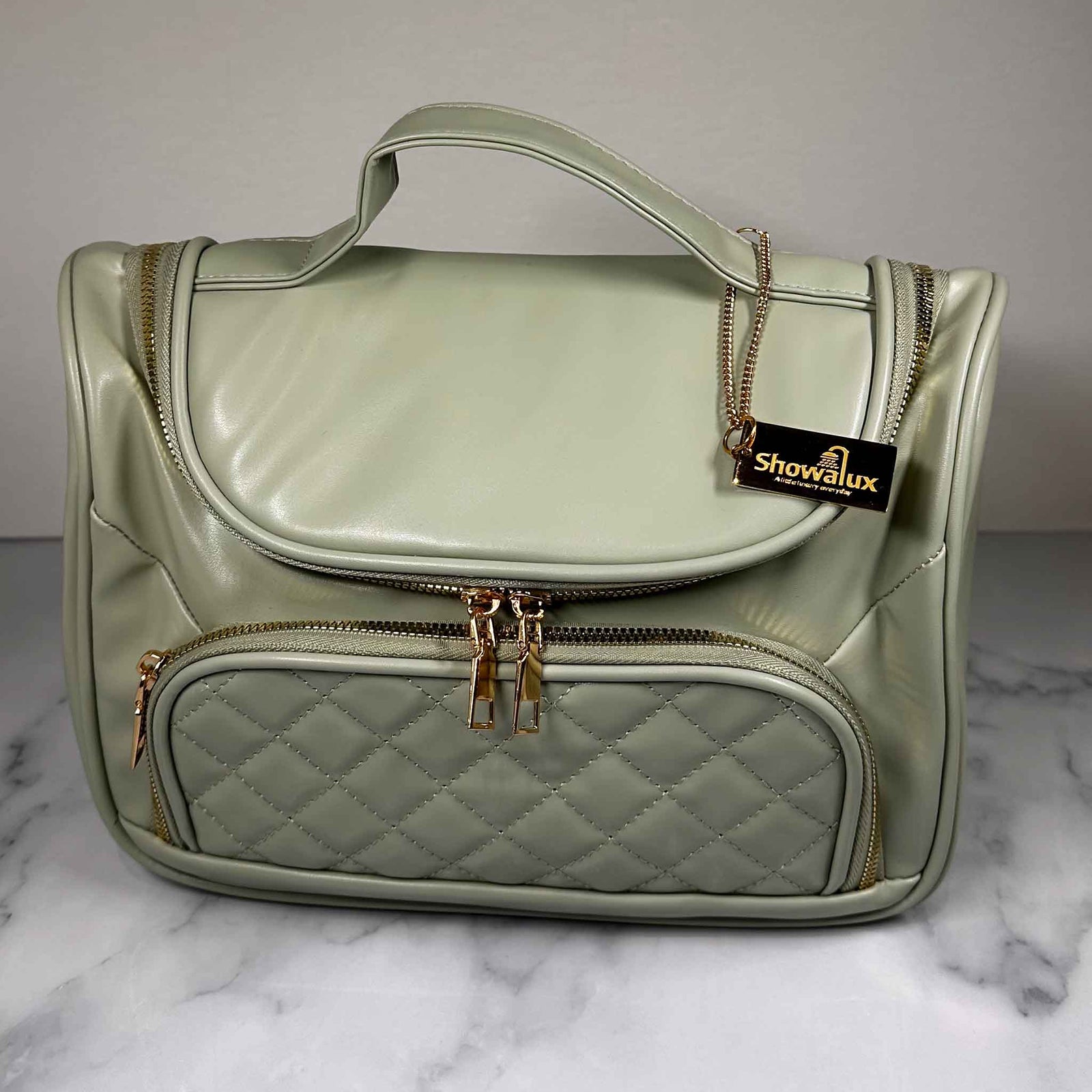 A green handbag style toiletry bag with duel zips for the main section, a side pouch and a gold tag with the showalux logo on a gold chain, on a marble top
