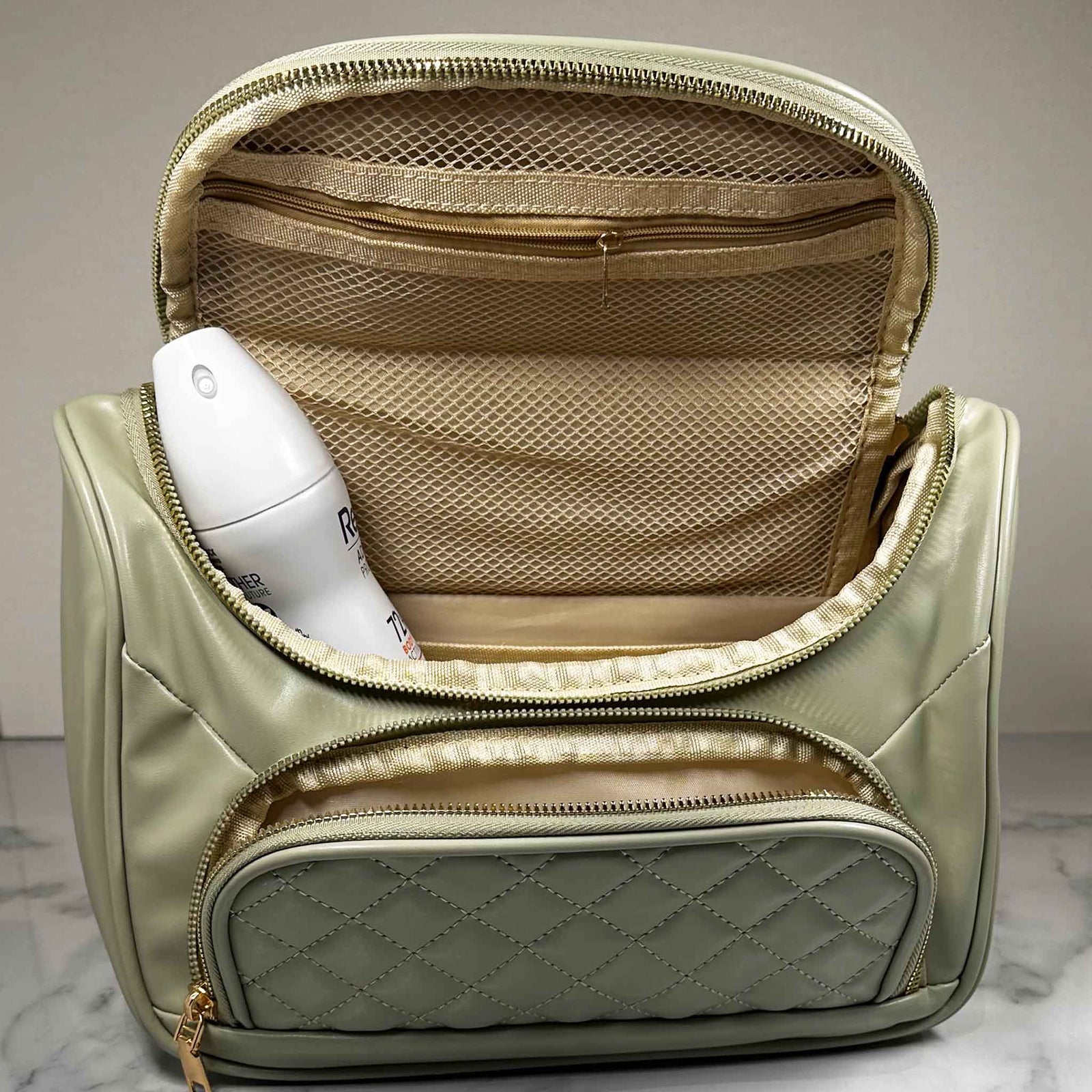A green handbag style toiletry bag with duel zips undone showing inside of main compartment with a tin of deodorant inside, a side pouch that is open on a marble top