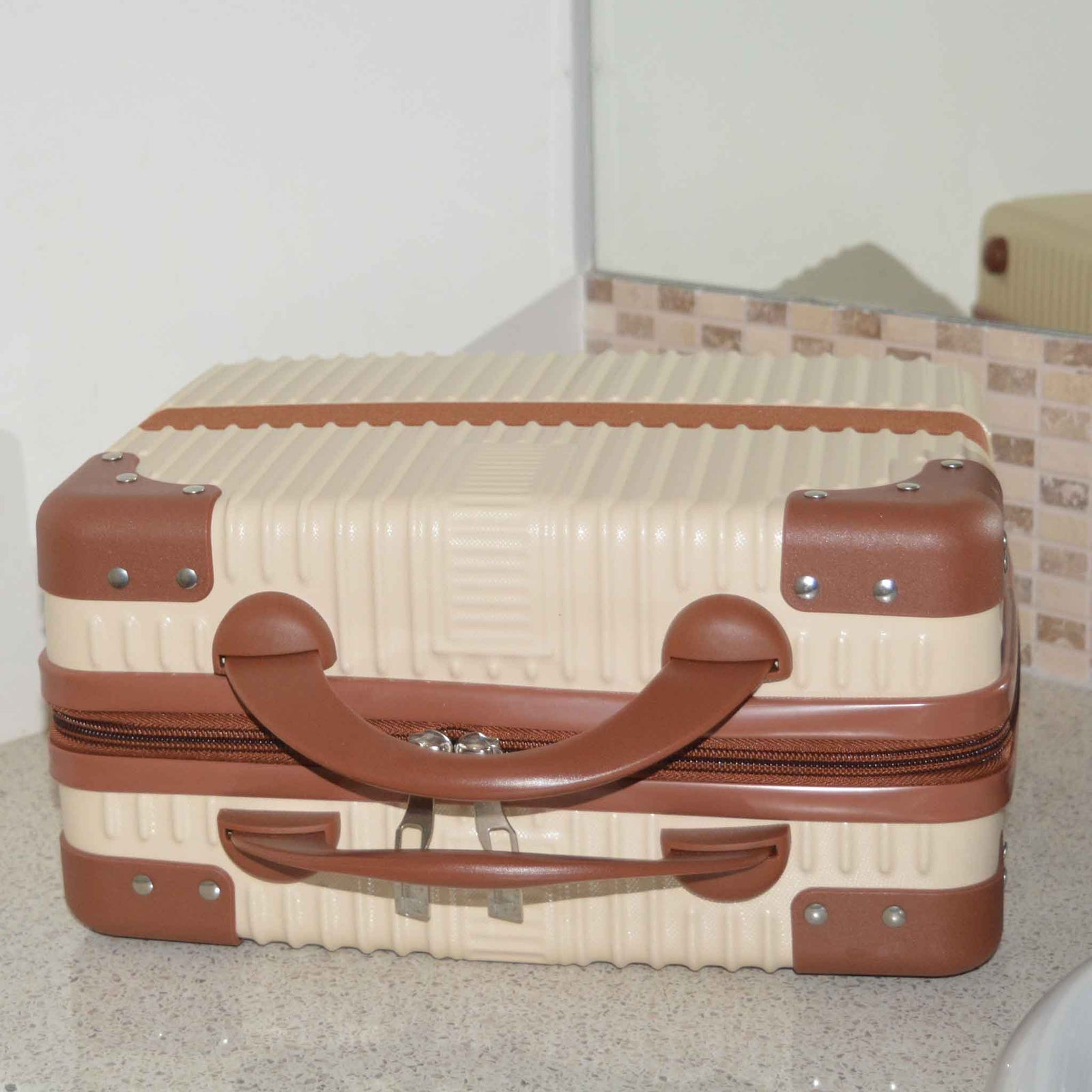 Suitcase Style Toiletry Bag closed and laying on its side on a bathroom bench. Beige case with brown corners, handles and strap