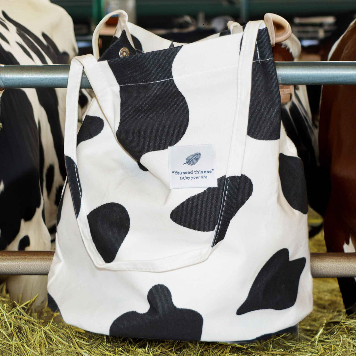 A corduroy tote bag in a black and white cow print sitting on a hay bale with cows in the background