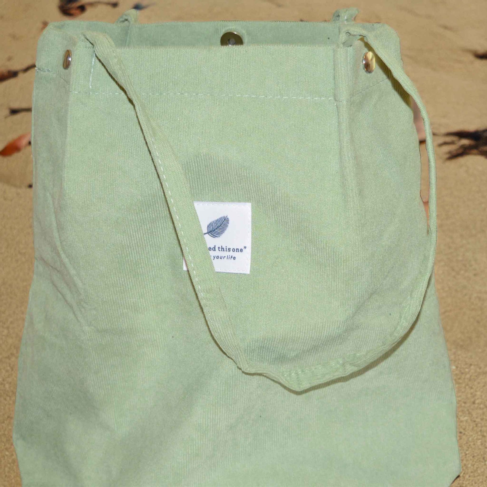 A light green corduroy tote bag with long handles on a background of sand 