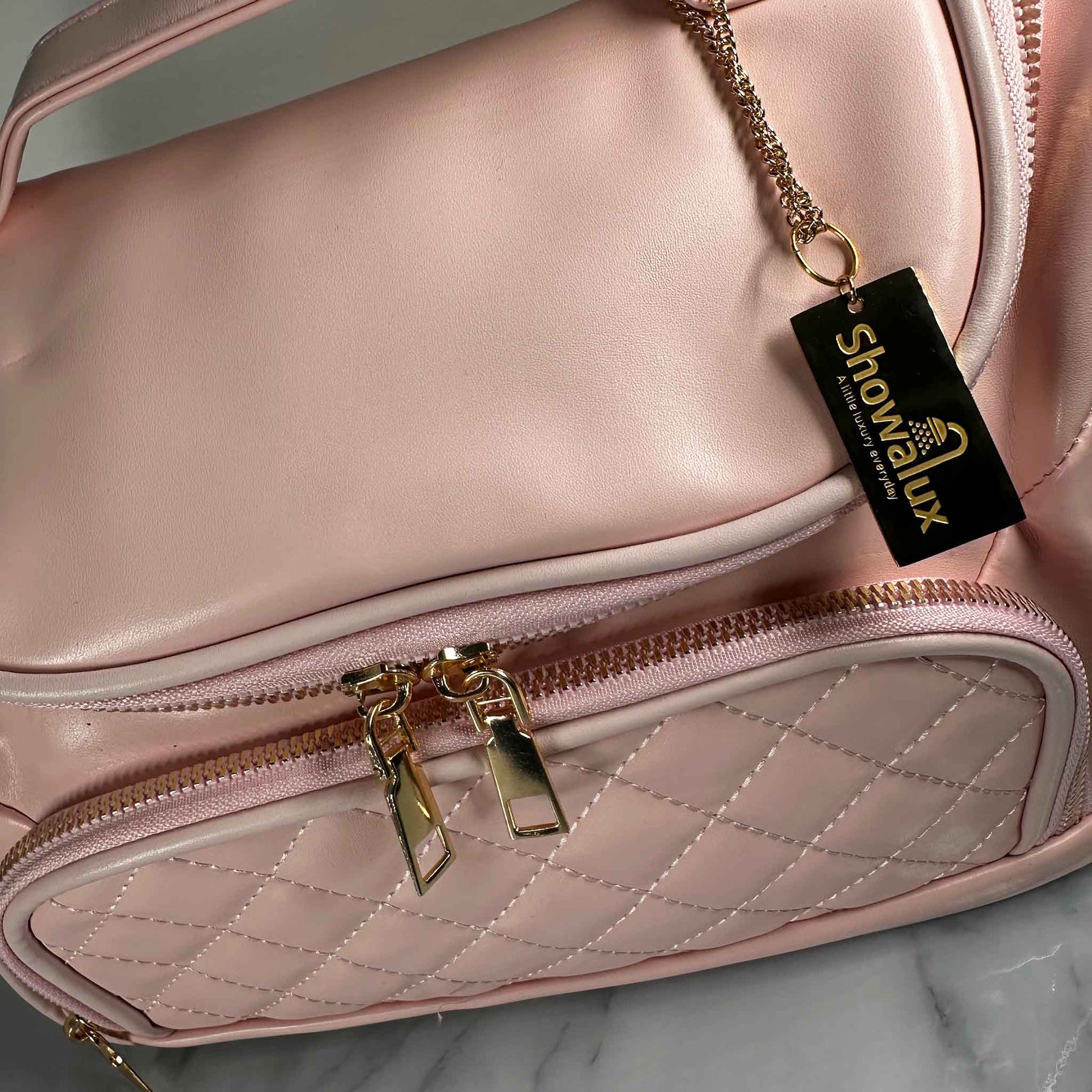 A close up of a pink handbag style toiletry bag with duel zips for the main section, a side pouch and a gold tag with the showalux logo on a gold chain, on a marble top