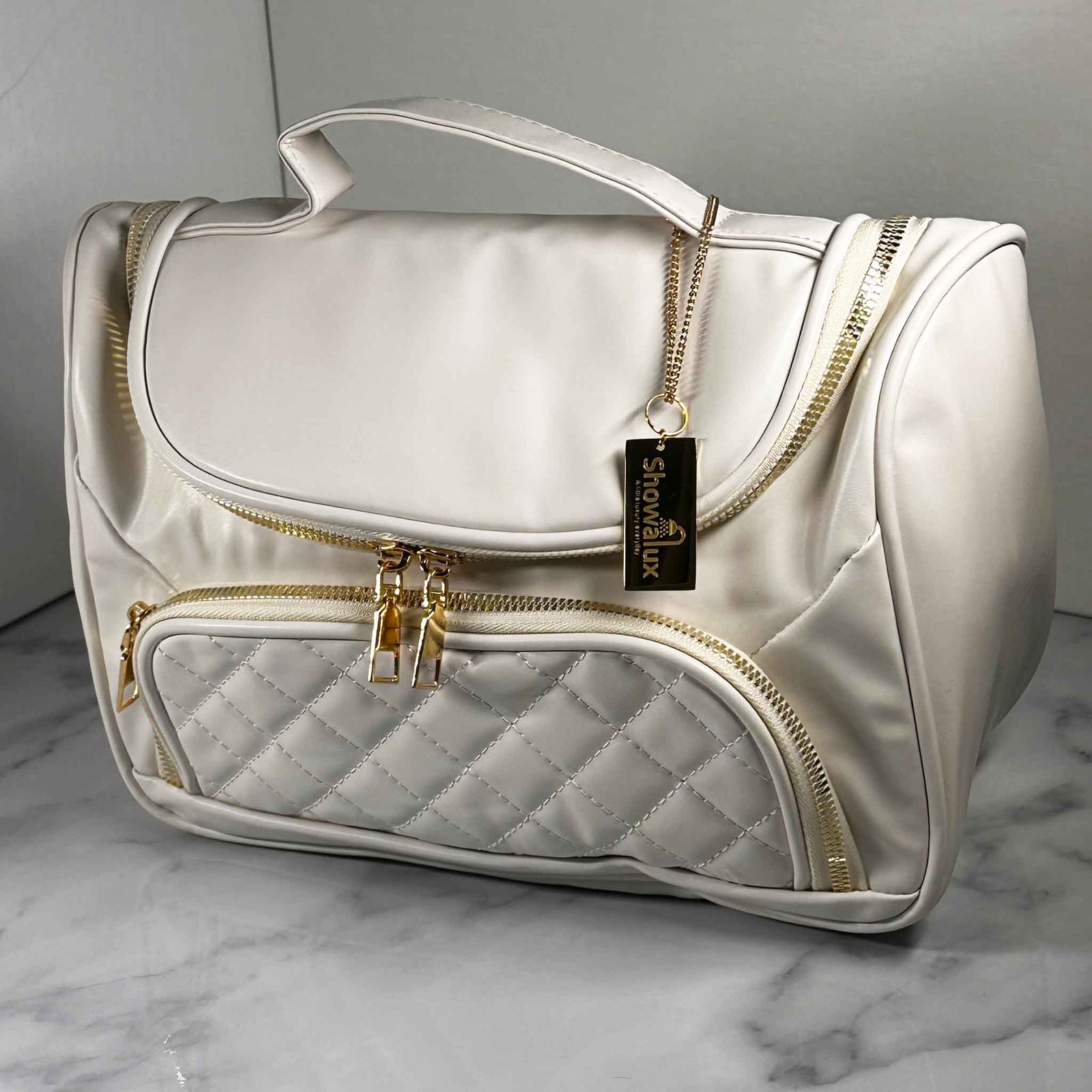 A white handbag style toiletry bag with duel zips for the main section, a side pouch and a gold tag with the showalux logo on a gold chain, on a marble top