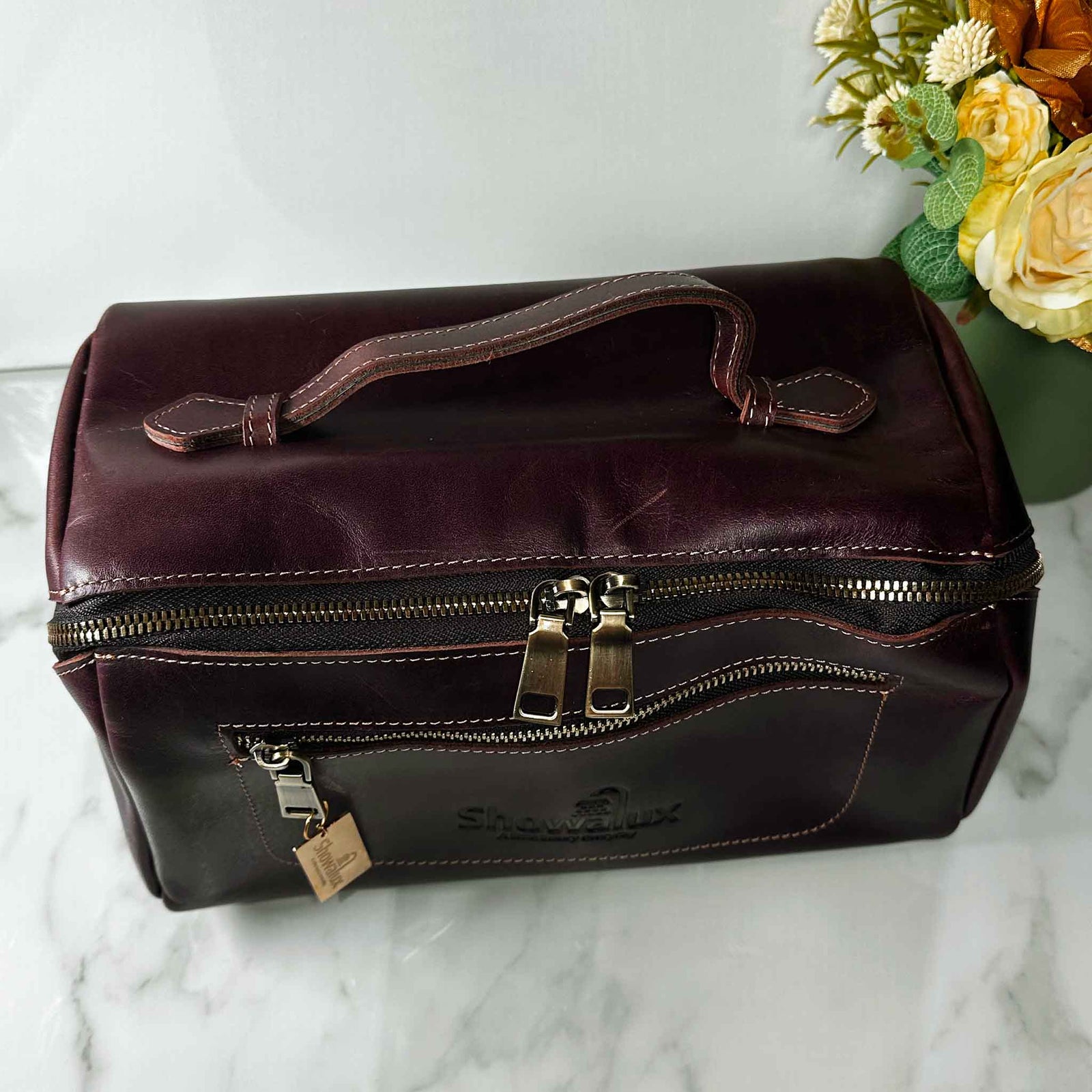 Brown leather toiletry bag with a visible brand logo on a marble surface with flowers in the background.