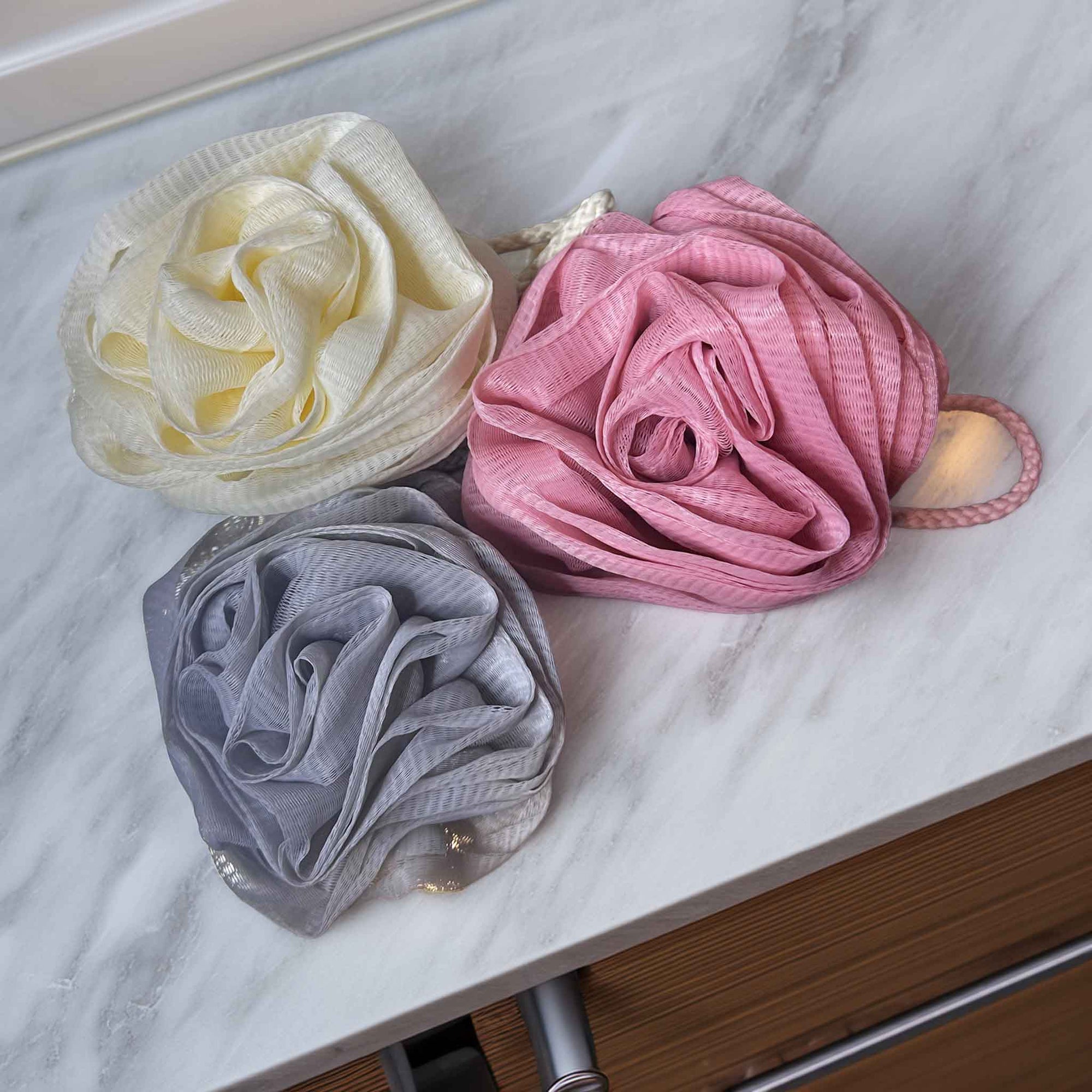 Three bath scrubs in white, pink, and gray on a marble surface.