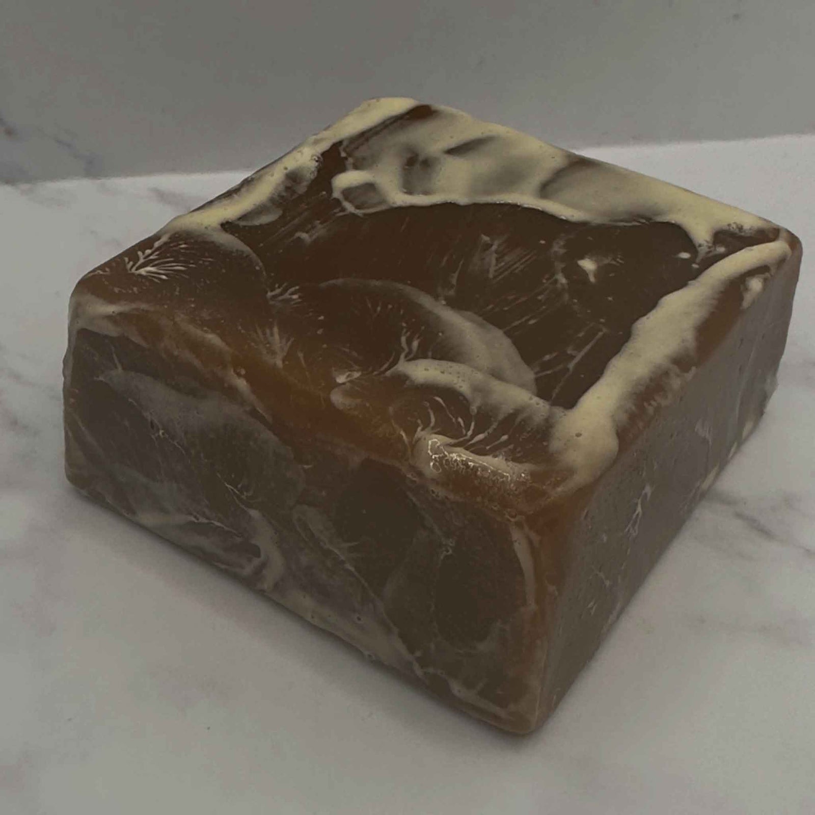 A dark brown soap on a flecked soap dish next to a soap wrapped in shrink wrap and labeled leaning against a pine cone.