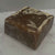 A dark brown soap on a flecked soap dish next to a soap wrapped in shrink wrap and labeled leaning against a pine cone.
