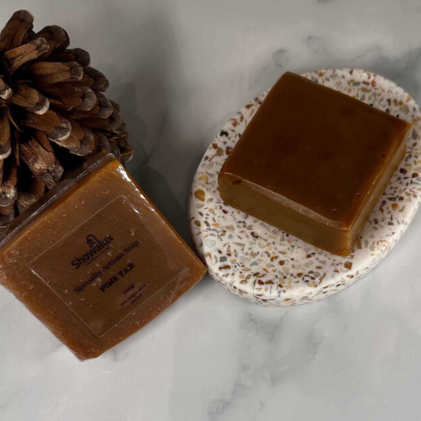A dark brown soap on a flecked soap dish next to a soap wrapped in shrink wrap and labeled leaning against a pine cone.
