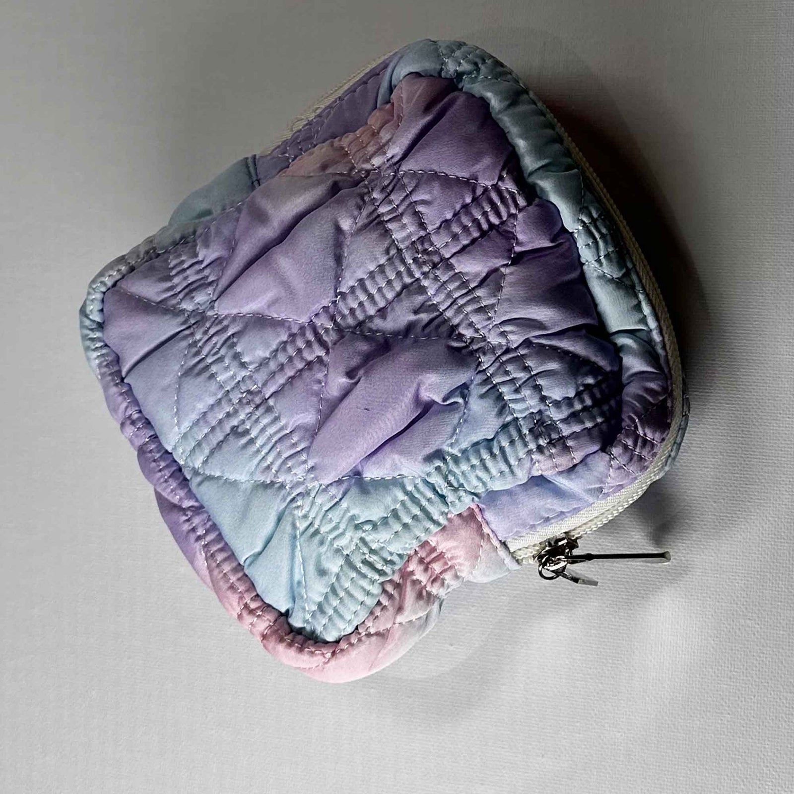 A square bag coloured with a mix of purple, blue and pink, zipped shut on a white background