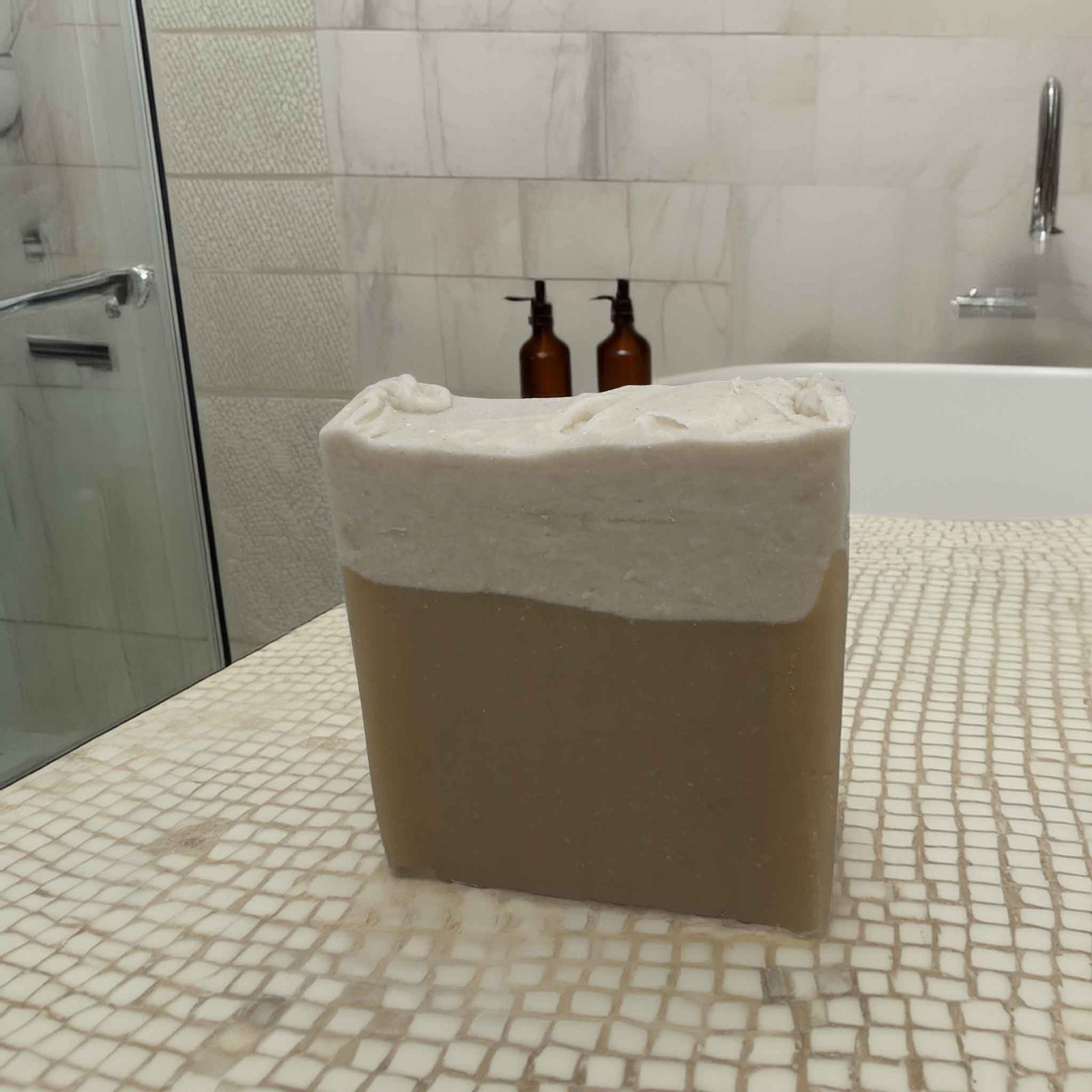 A square bar of soap that is white on top and natural colour on the bottom 2/3s on a tiled bathroom bench