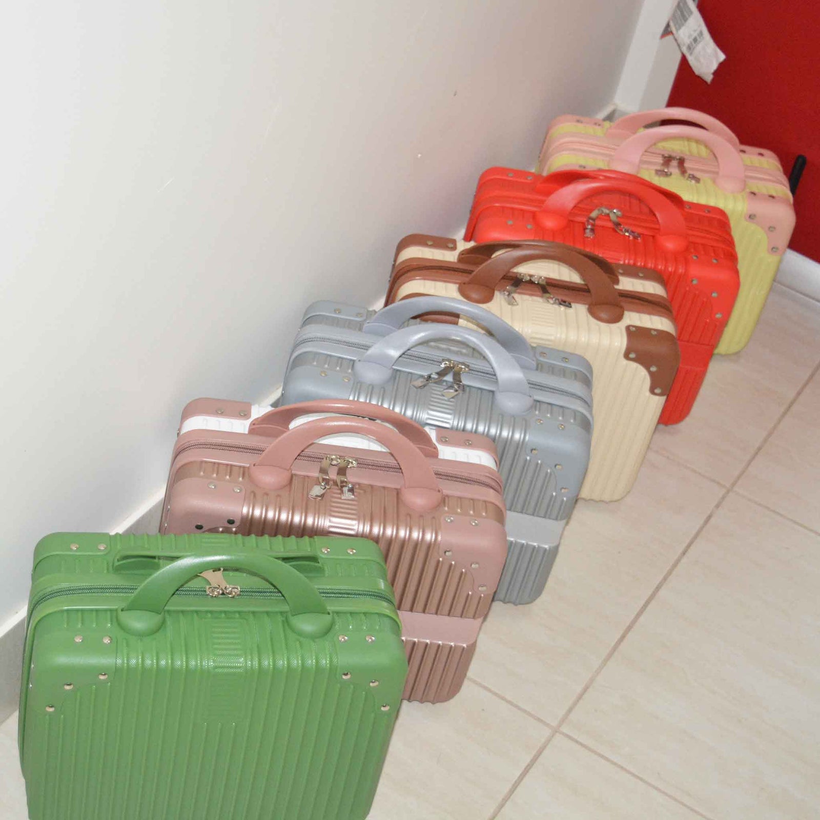 Green, rose gold, silver, cream, red and cream suitcase bags lined up on the ground along a white wall with a large case in the background