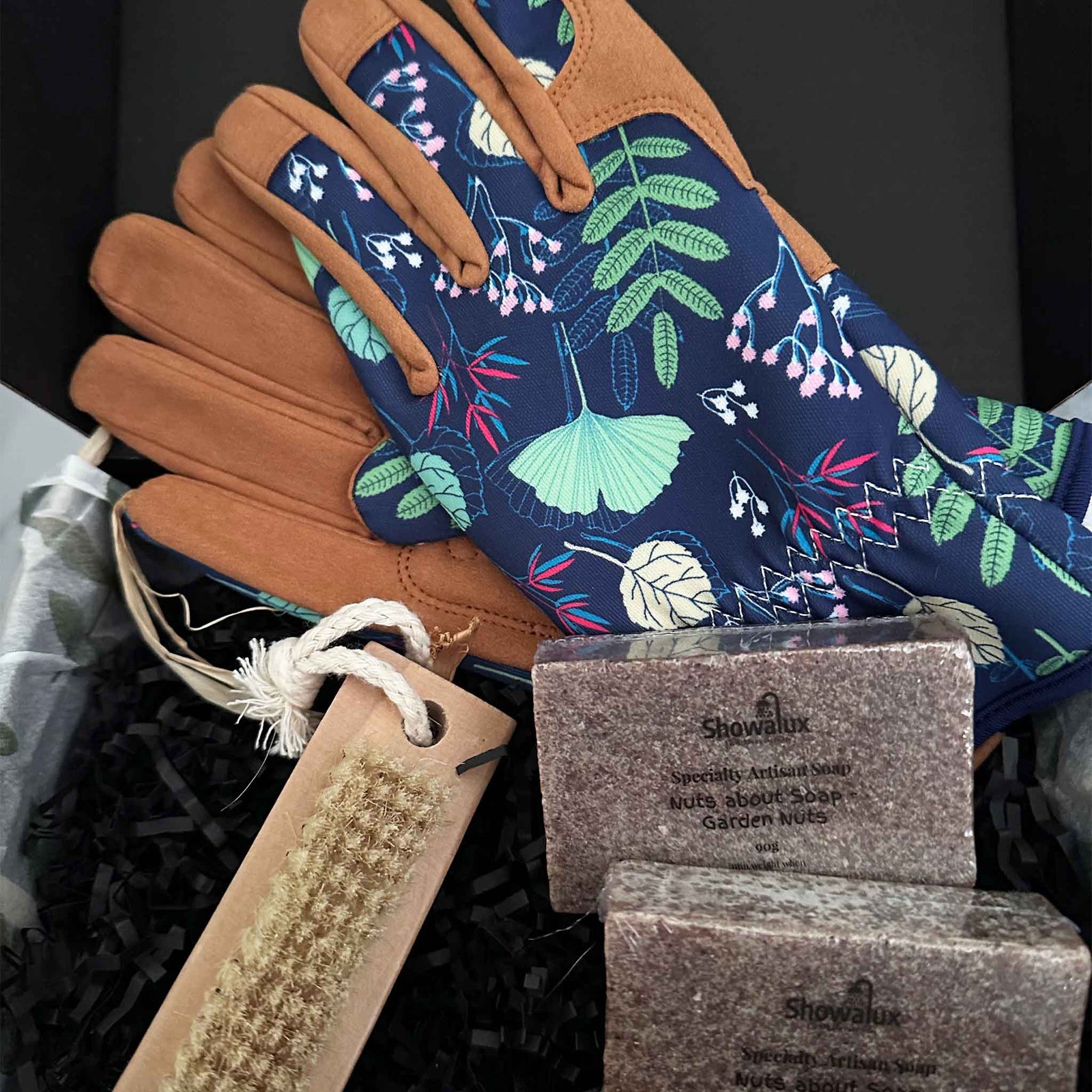 Pair of gardening gloves with floral pattern and brown leather accents, along with two Showalux soaps and a brush on a black background.