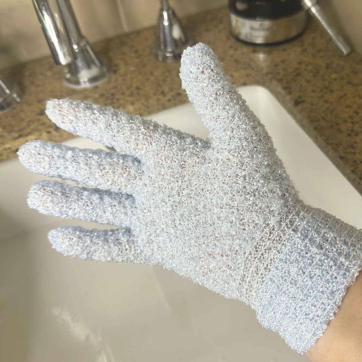 Blue exfoliating glove on hand with sink in the background
