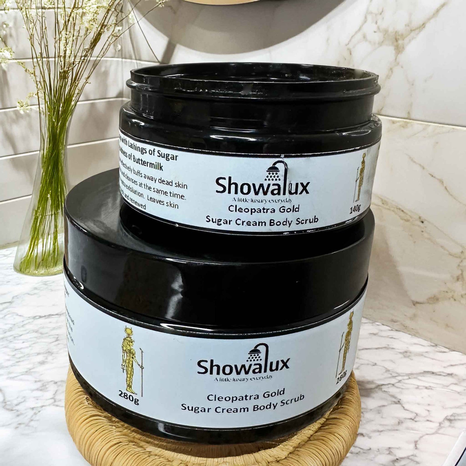 Two Showalux Cleopatra Gold Sugar Cream Body Scrubs stacked on a marble surface with a vase in the background.