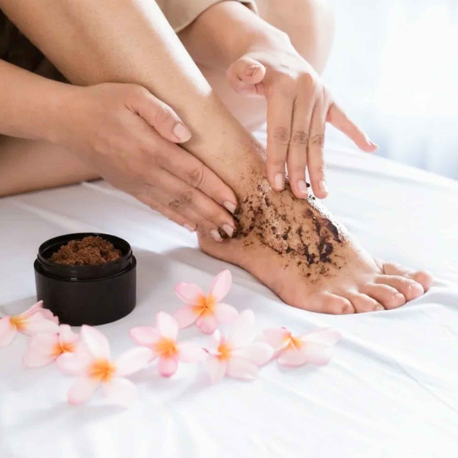 Person applying a brown scrub to their foot with flowers and a container of scrub on a white surface.