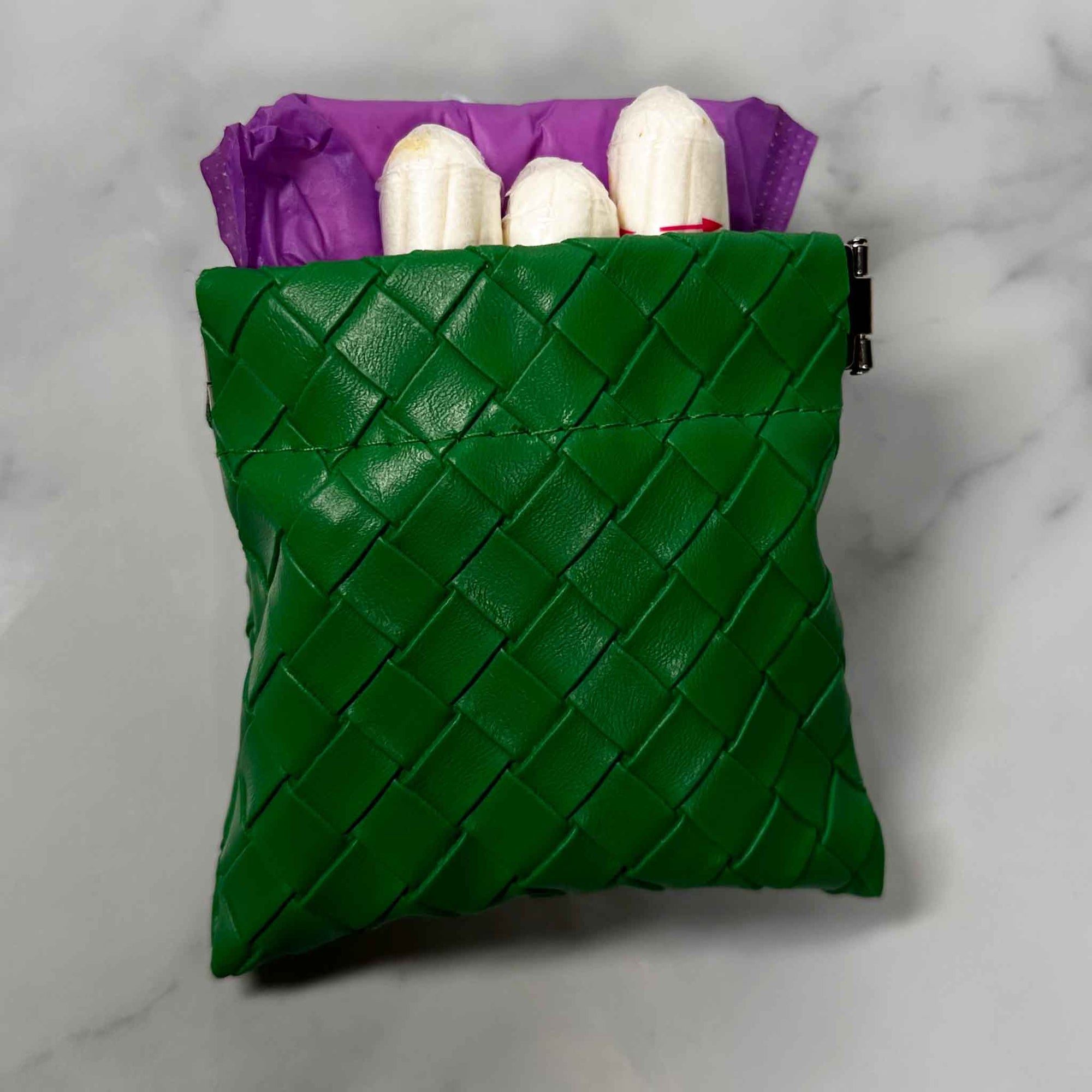 Green woven Showalux personal bag showing contents of  purple panty liners and tampons on a marble surface